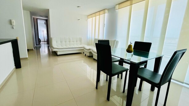A modern living room with a glass dining table, black chairs, a white sofa, and a large window with blinds. - 3-bedroom apartment in enjoyable Barranquilla with AC for relaxing stays (Barranquilla)