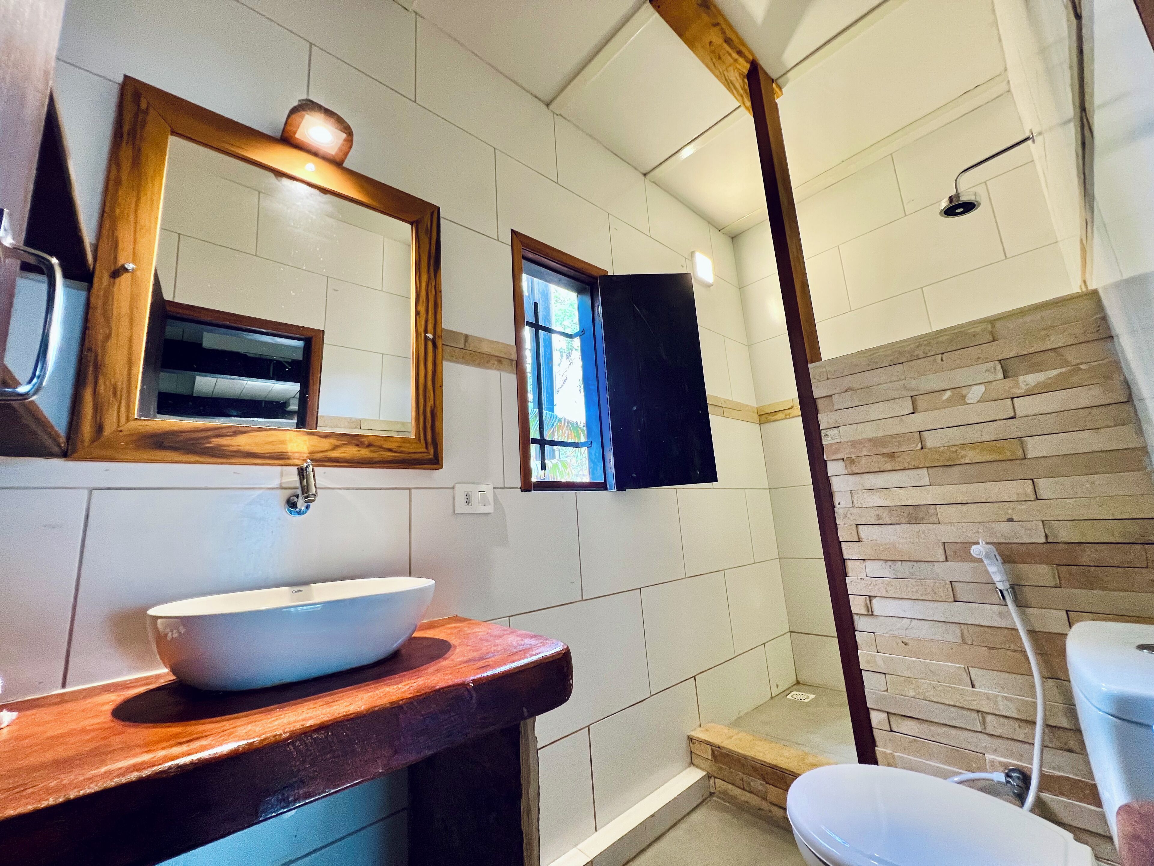A bathroom with a wooden vanity, a large mirror, a white sink, a shower with a stone wall, and a window.