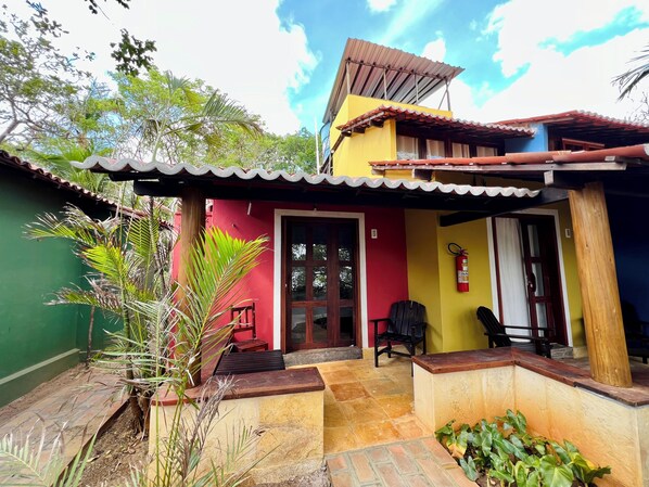 Ein zweigeschossiges Haus mit roter und gelber Fassade, einer gefliesten Terrasse und einer kleinen Veranda mit einem Stuhl.