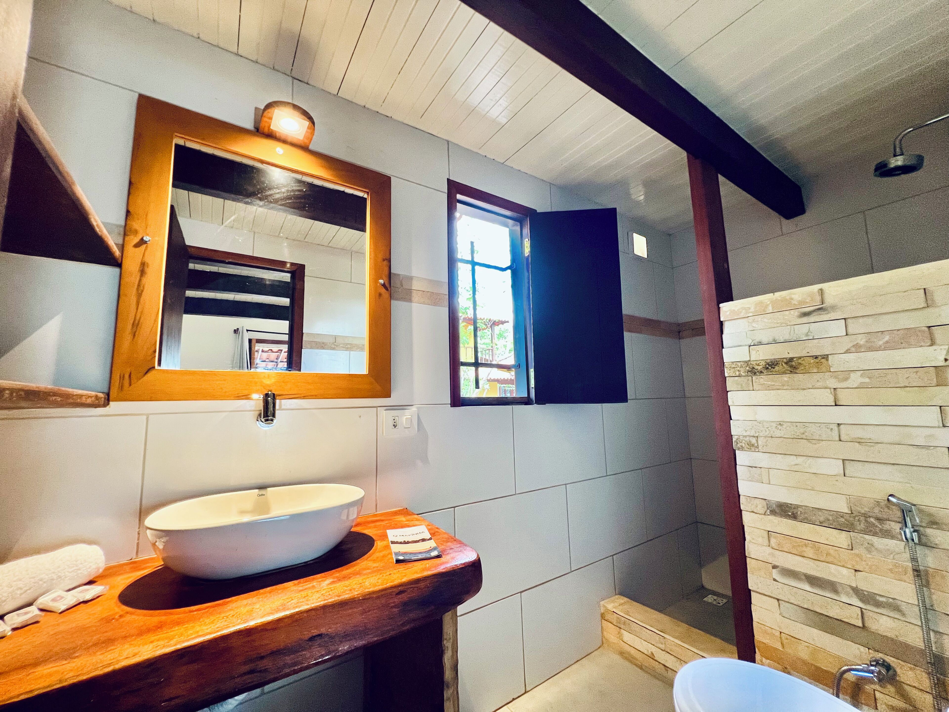Un baño con tocador de madera, un espejo grande, un lavamanos blanco, ducha con pared de piedra y ventana con vistas a follaje.