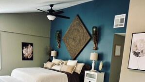 A bedroom with a bed, a nightstand, a chair, a ceiling fan, and a decorative wall piece.
