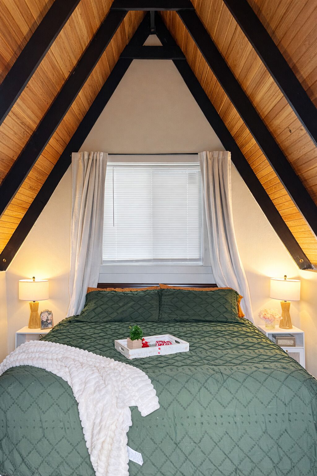 Un dormitorio con techo inclinado, una cama con cubrecama acolchado verde, una manta blanca y una mesita de noche con lámpara y marco de foto.