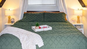 Quarto com teto inclinado, cama com colcha verde acolchoada, manta branca e mesa de cabeceira com abajur e moldura de quadro.