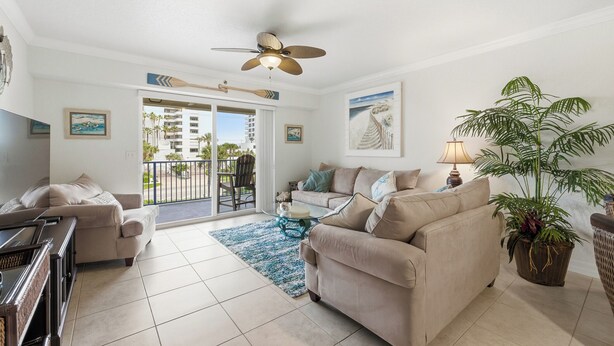 A spacious living room with a beige sofa, a glass coffee table, and a ceiling fan. There is a large window with a view of palm trees and a building. - Oceanwalk 3BR | Ocean Views, Private Balcony & Steps to Car-Free Beach (New Smyrna Beach)