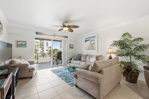 Ein geräumiges Wohnzimmer mit einem beigen Sofa, einem Glastisch und einem Deckenventilator. Ein großes Fenster bietet Blick auf Palmen und ein Gebäude.