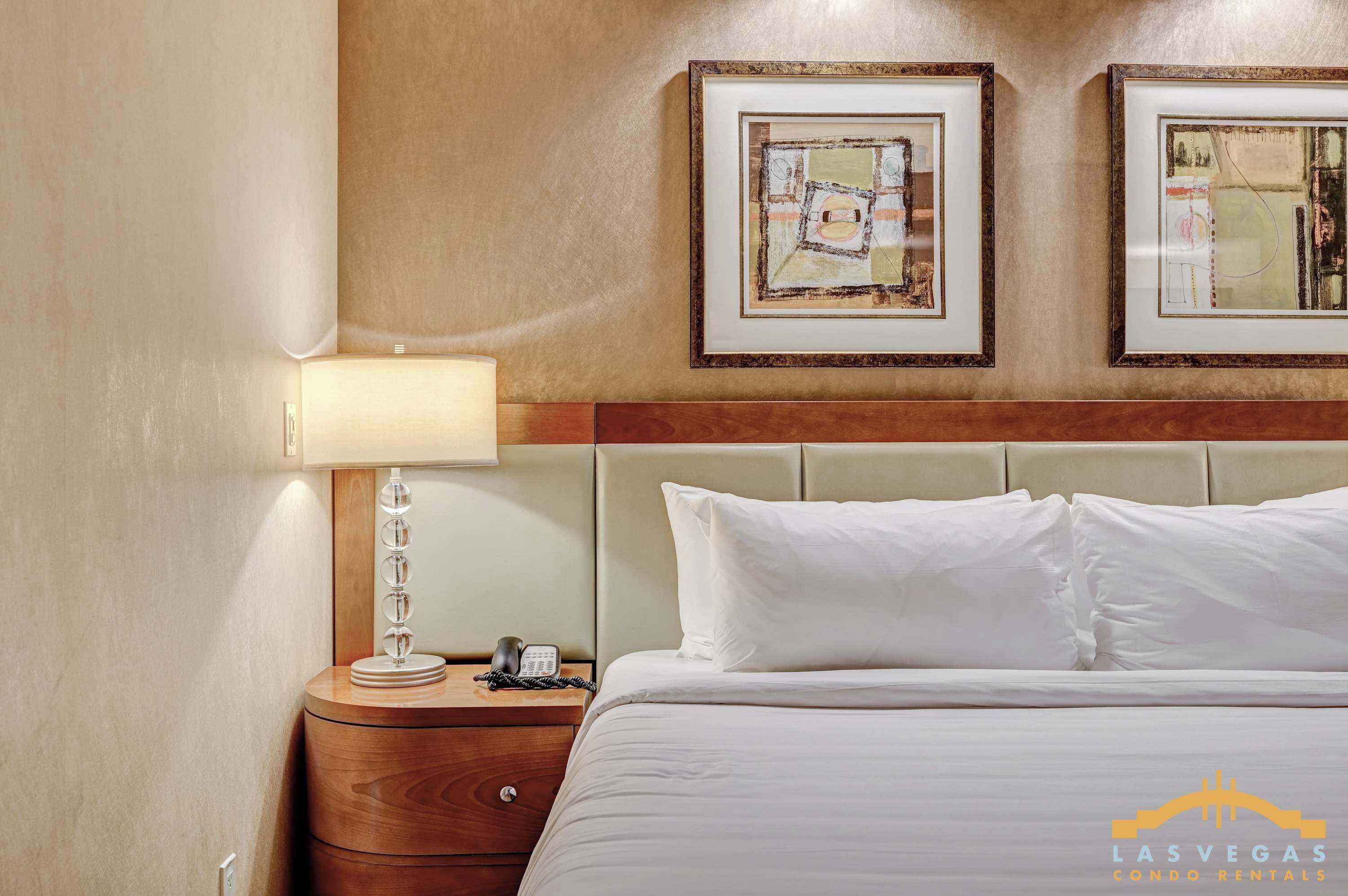 Una habitación de hotel con una cama, una lámpara de noche, una mesita de noche con un teléfono y dos cuadros en la pared.