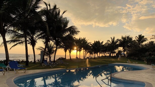 Ein Schwimmbecken mit Liegestühlen, Palmen und Blick auf den Ozean bei Sonnenuntergang.