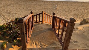 Eine Holztreppe führt zu einem Sandstrand, im Hintergrund sind Meereswellen zu sehen.
