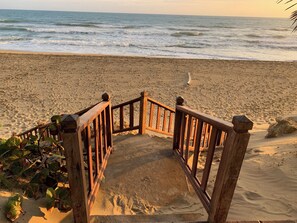 Eine Holztreppe führt zu einem Sandstrand, im Hintergrund sind Meereswellen zu sehen.