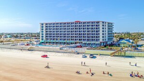 Ein mehrstöckiges Hotel mit Balkonen, in Strandnähe mit sandigem Ufer und einigen parkenden Autos.