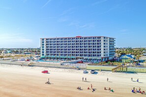 A multi-story hotel with balconies, located near a beach with a sandy shore and a few parked cars. - Upgraded Beachfront Condo w/ Balcony & King Bed | Walk to Boardwalk (Daytona Beach)