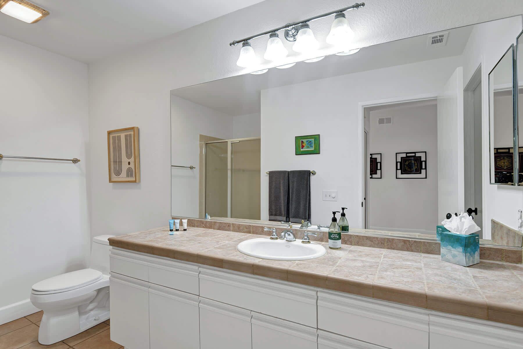 A modern bathroom with a white toilet, a beige countertop, a large mirror, and a shower area.