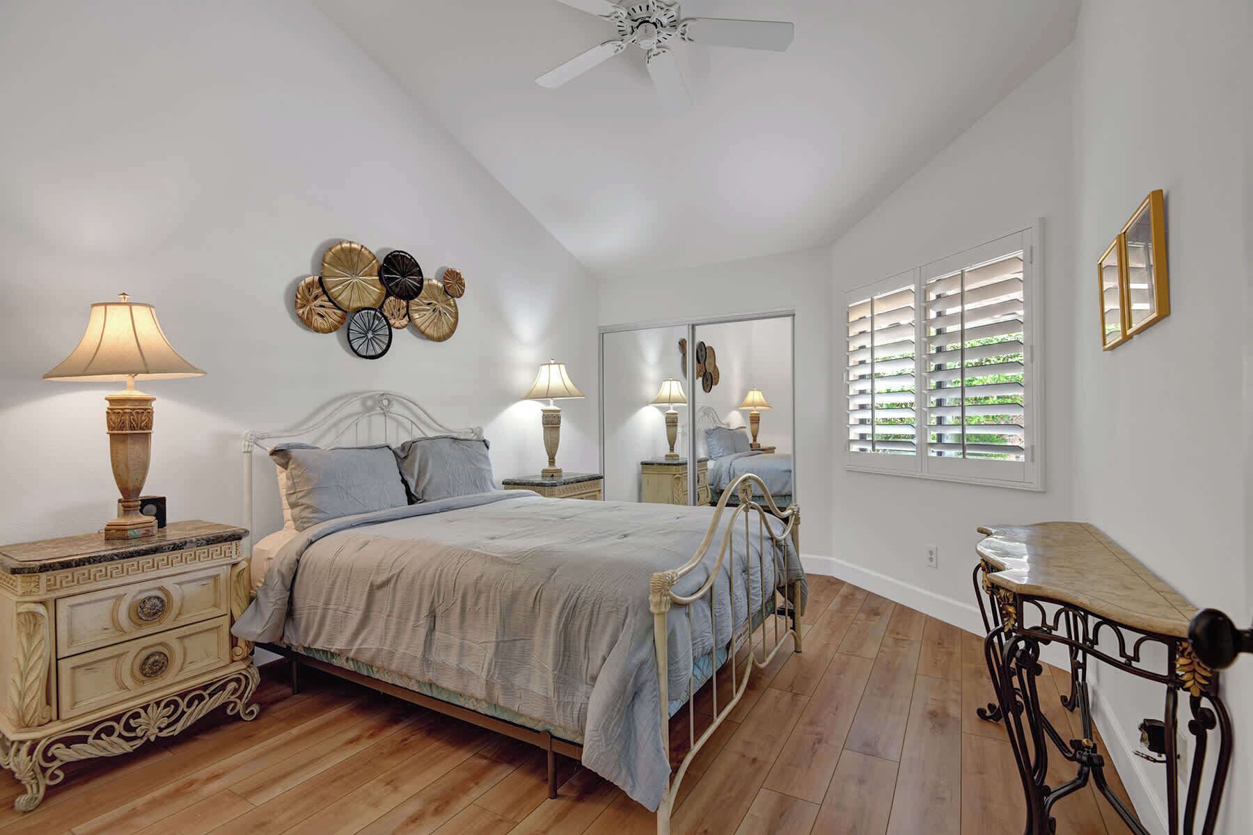 Une chambre à coucher comprenant un lit, deux tables de chevet, un miroir, un ventilateur de plafond et une fenêtre avec des volets.