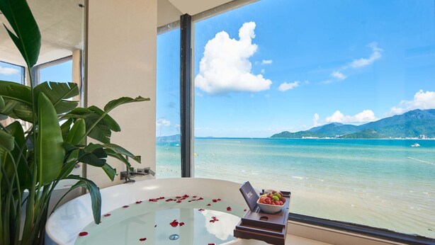 A bathroom with a bathtub filled with water and rose petals, a bowl of fruit, and a view of a beach and mountains through large windows. - Santa Monica Seaview Vacation Home (Shenzhen)