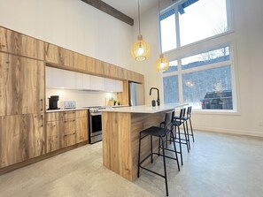 A modern kitchen with wooden cabinets, a central island, bar stools, and large windows. - Luxury nature chalet | Modern | Spa (St-Raymond)
