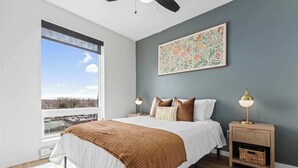 Une chambre avec une grande fenêtre, un lit recouvert d’une literie blanche, un plaid marron, deux tables de chevet avec des lampes et une œuvre d’art accrochée au mur.