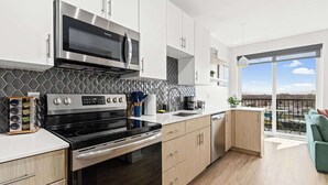 Une cuisine moderne dotée d’appareils électroménagers en acier inoxydable, d’un four à micro-ondes et d’une cuisinière. Elle se distingue par ses meubles de couleur bois clair, son plan de travail noir et sa vue sur l’extérieur grâce à de grandes fenêtres.