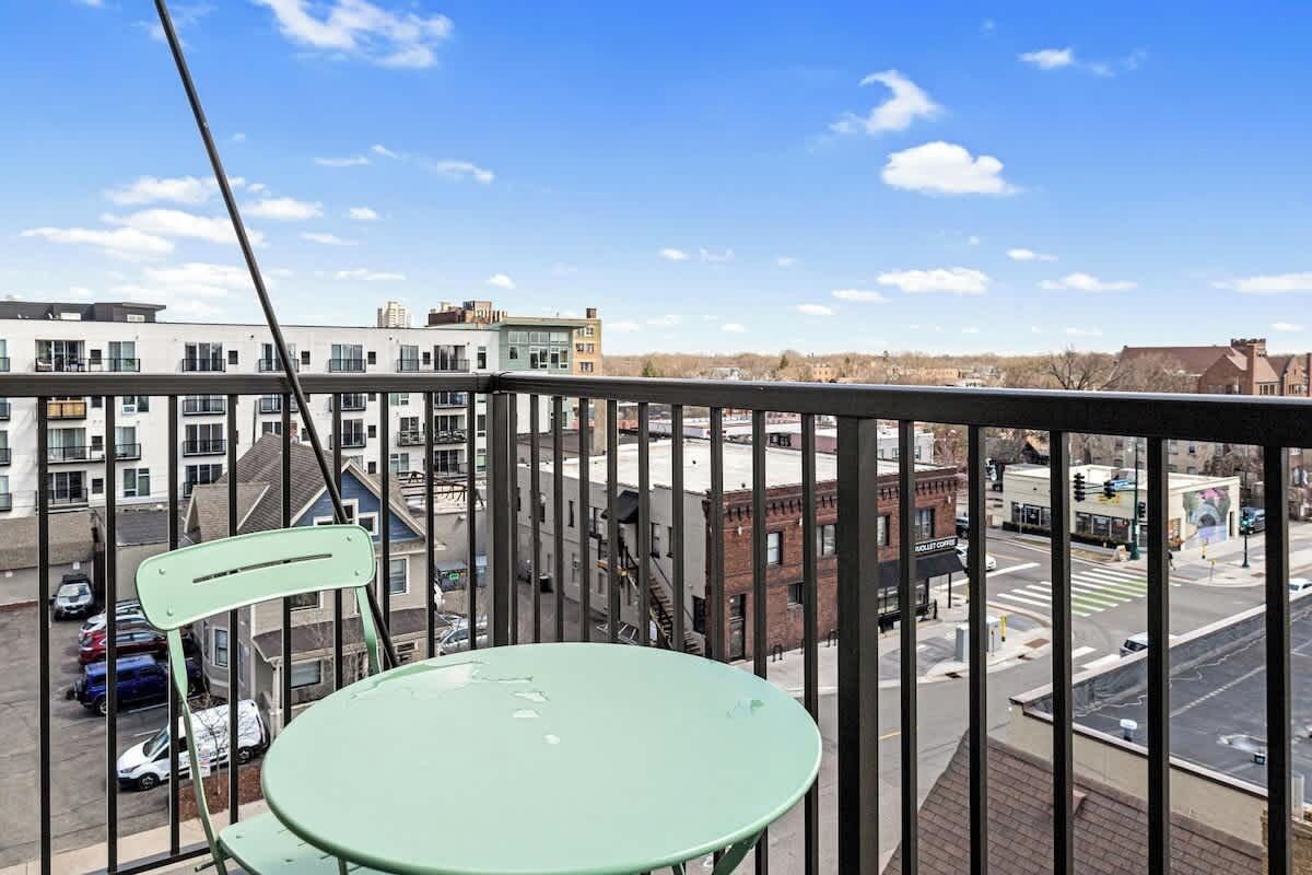 A balcony with a green chair and a table, overlooking a street with buildings and a clear sky.
