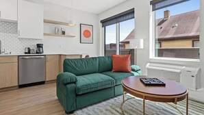 Eine moderne Wohnung mit einem grünen Sofa, einem runden Holztisch und einer Küchenzeile inklusive Geschirrspüler und Kaffeemaschine.
