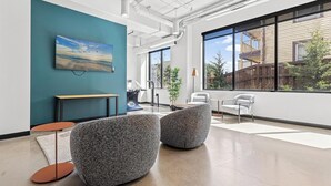 Ein moderner Arbeitsraum mit einer türkisfarbenen Akzentwand, einem großen Fenster mit Blick auf Gebäude und Bäume sowie zeitgemäßen Möbeln, darunter zwei graue Sitzsäcke und ein kleiner runder Tisch.