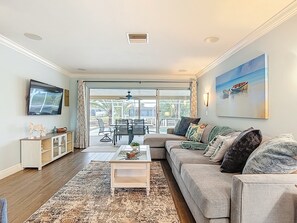 A living room with a sofa, coffee table, TV, and a view to the outside. - Heated Saltwater Pool, 3 Bed, 2 Bath Joyful Waterside (Cape Coral)