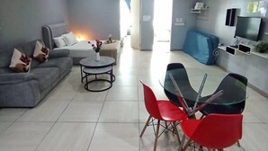 Een moderne woonkamer met een grijze bank, een ronde salontafel en rode stoelen. Er is een plafondventilator en een airconditioning.