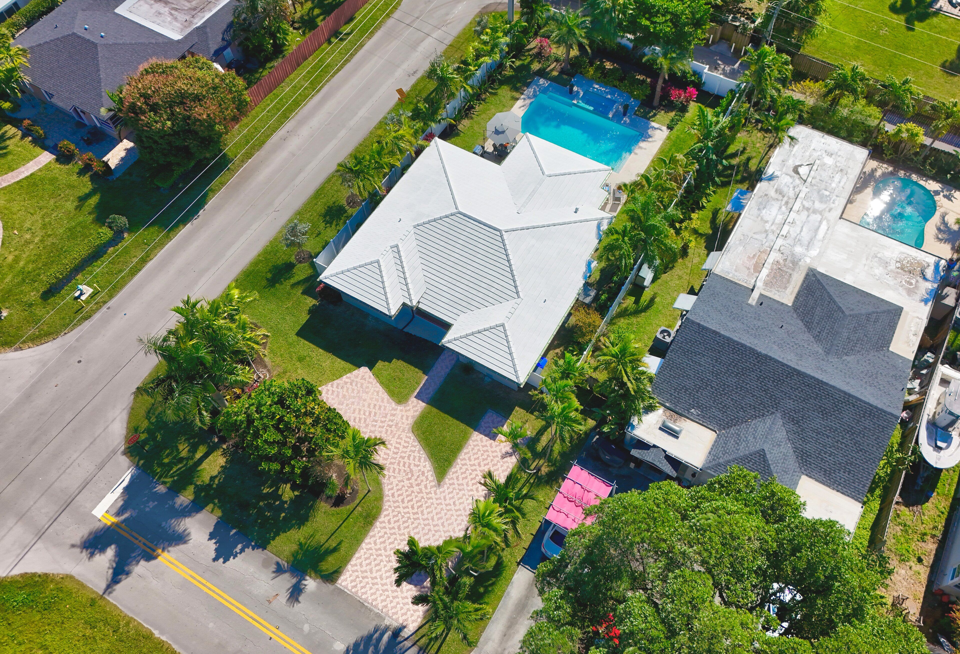 Eine Vogelperspektive zeigt eine Wohngegend mit einem Haus, das ein großes, weißes, geometrisches Dach, einen Swimmingpool und viel Grün hat.