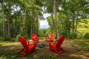 A forest with four red Adirondack chairs arranged in a circle around a campfire. - Luxury 4 BR Mountain Escape: Views, Hot Tub & Pool Table (Banner Elk)