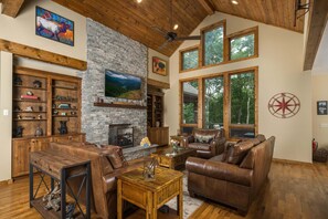 A spacious living room with a stone fireplace, leather furniture, and large windows offering a view of the outdoors. - Luxury 4 BR Mountain Escape: Views, Hot Tub & Pool Table (Banner Elk)