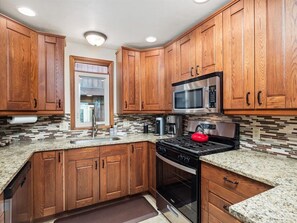 A kitchen with wooden cabinets, granite countertops, a stainless steel microwave, a red kettle, and a built-in oven. - Raven's Call Chalet, 6BR in Downtown Breck! (Breckenridge)