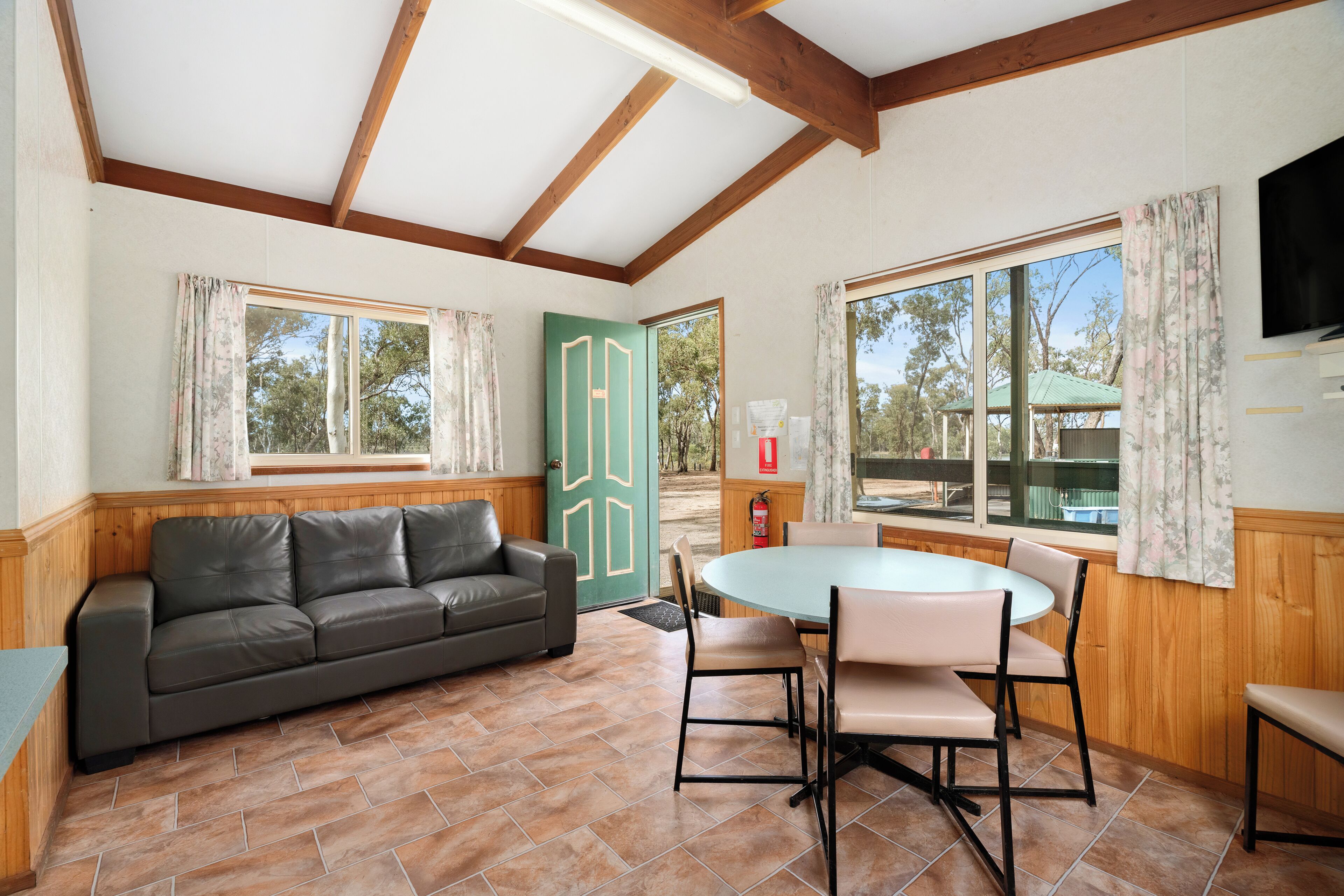A room with a wooden ceiling, a green door, a sofa, a table with chairs, and large windows.