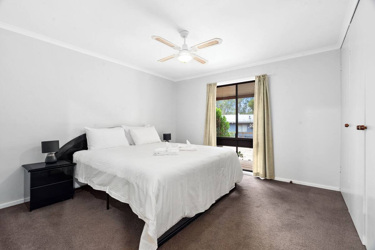 A bedroom with a bed, nightstand, lamp, ceiling fan, and a window with curtains.