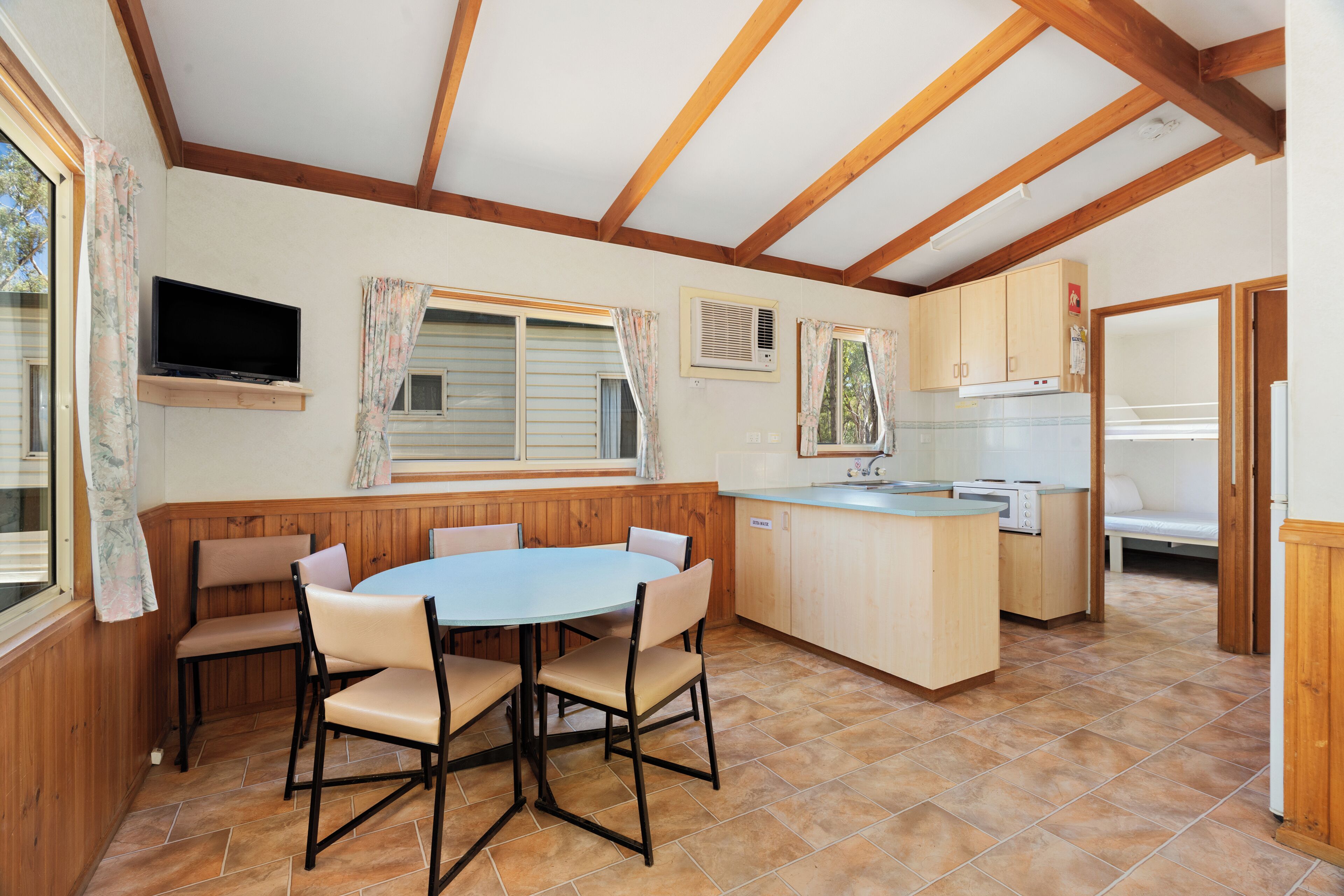 A kitchen with wooden cabinets, a dining table with chairs, a built-in TV, and a window with curtains.