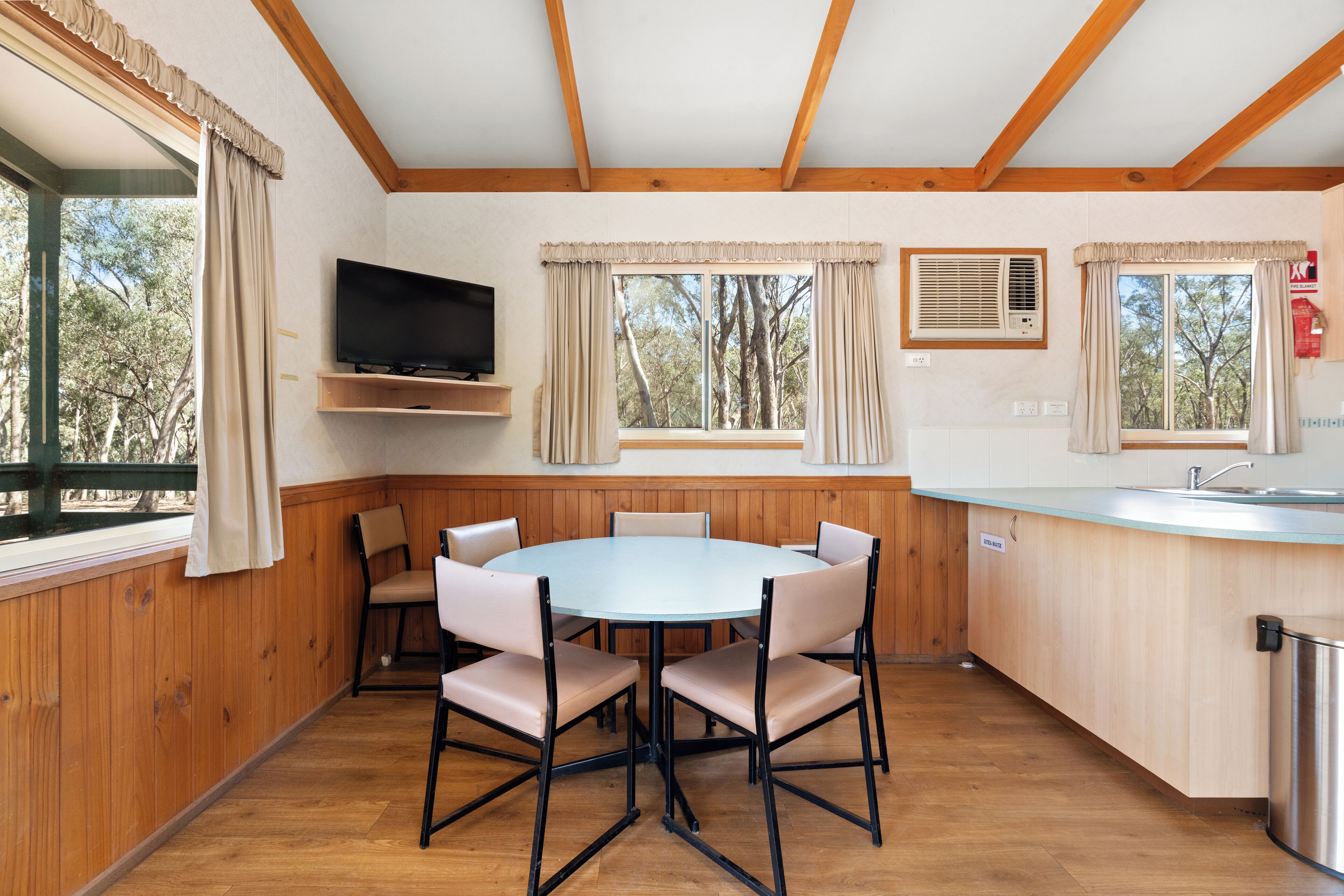A room with a round table and chairs, a television, a window with curtains, a sink, and a fire extinguisher.