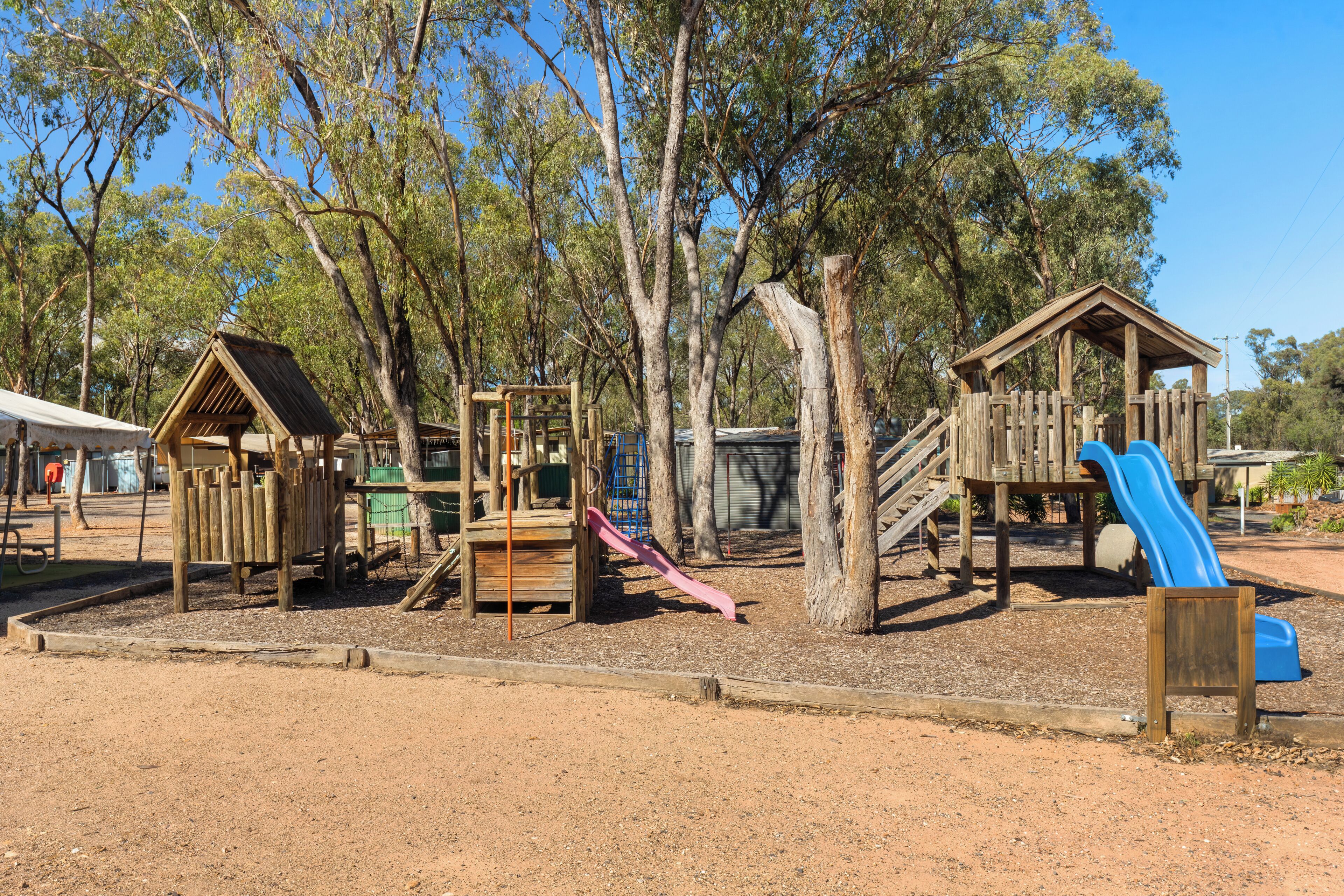 A playground with wooden structures, including a slide and climbing frames, surrounded by trees.