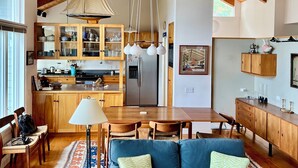A spacious living area with a wooden ceiling, a blue sofa, a dining table, and a kitchen with wooden cabinets and a refrigerator. - Mid-coast Dream- Beautiful Oceanfront home with spectacular views! (St. George)