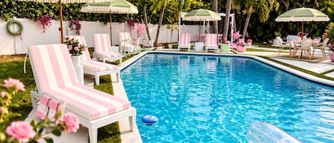 A swimming pool with lounge chairs and umbrellas, surrounded by palm trees and flowers.