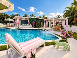 A swimming pool with lounge chairs, a covered patio area, and a small building with a green wall.