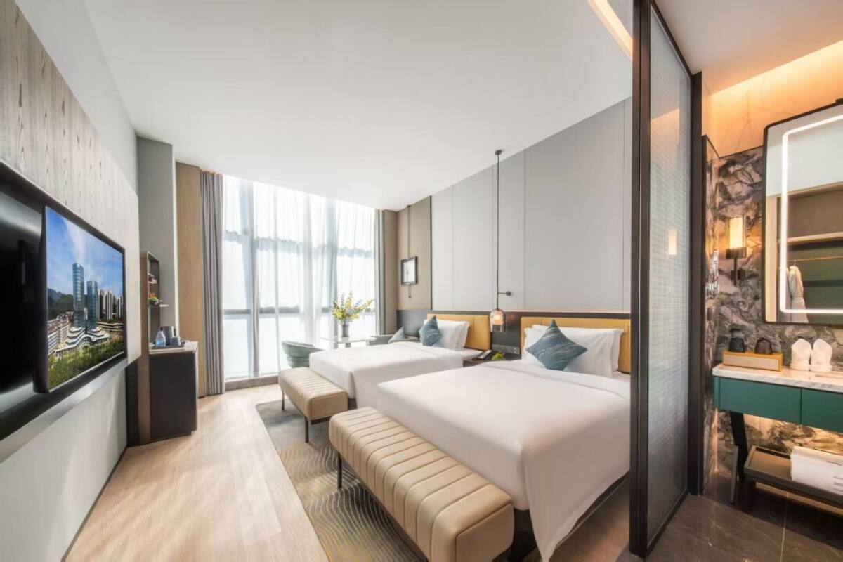 A hotel room with two beds, a TV, a desk, and a bathroom with a mirror and sink.