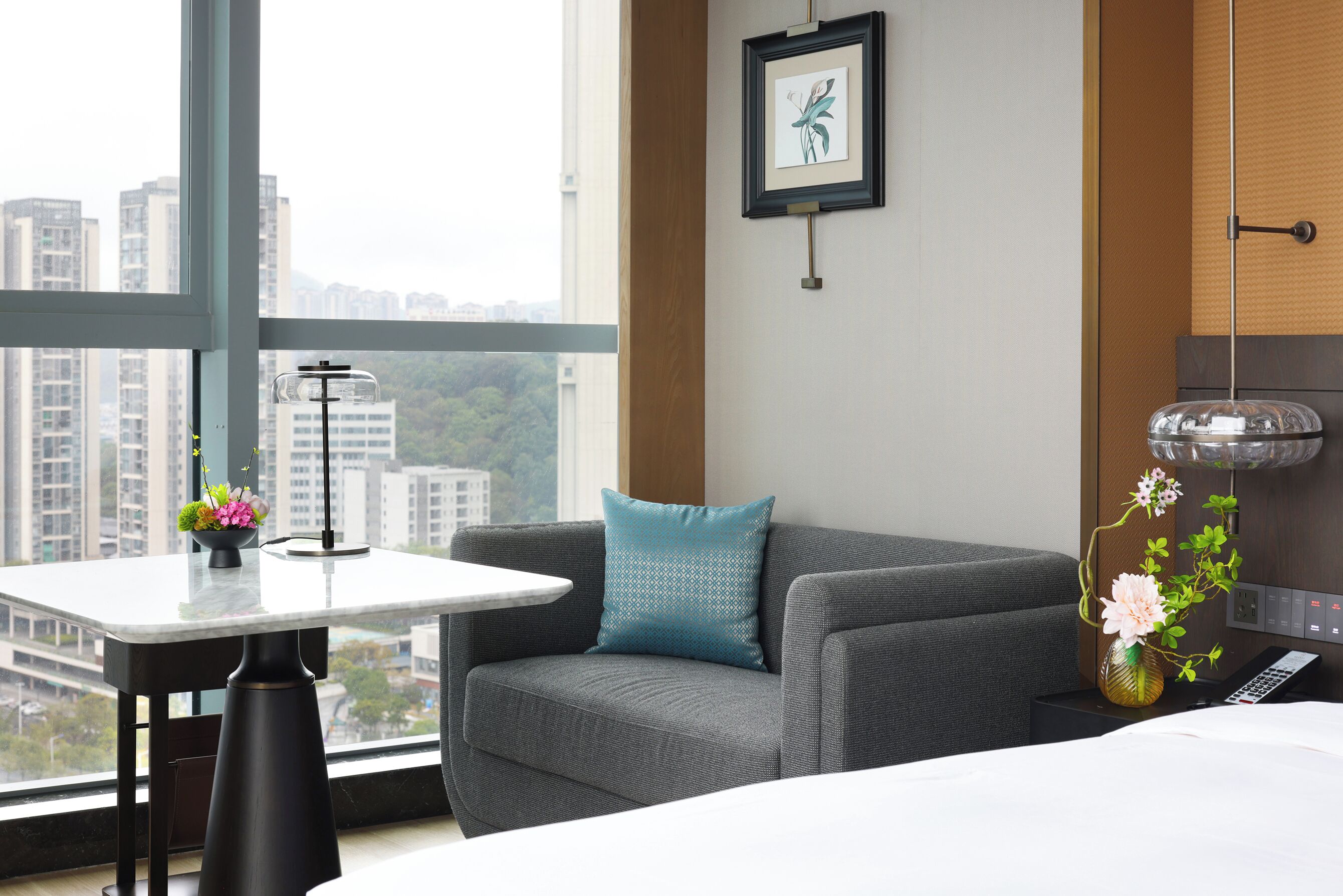 Quarto de hotel com um sofá cinzento, uma mesa pequena com um vaso de flores, um quadro na parede e vista para edifícios altos pela janela.