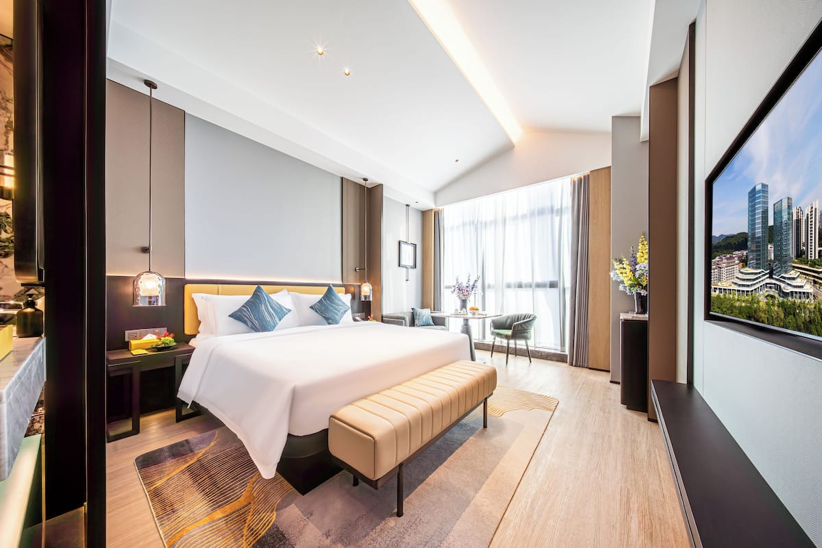 A modern hotel room with a large bed, a bench, a desk, and a large window offering a city view.