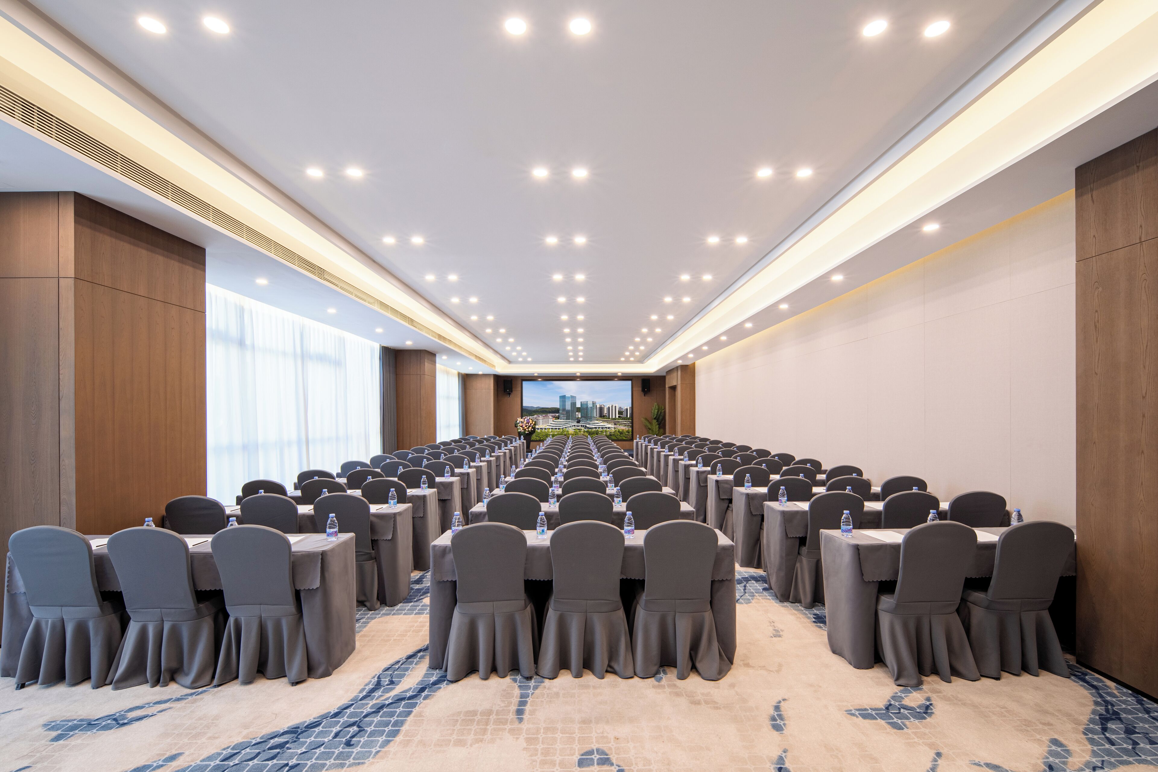 Un salón de conferencias con filas de mesas y sillas, suelo alfombrado y vistas a la ciudad a través de las ventanas.