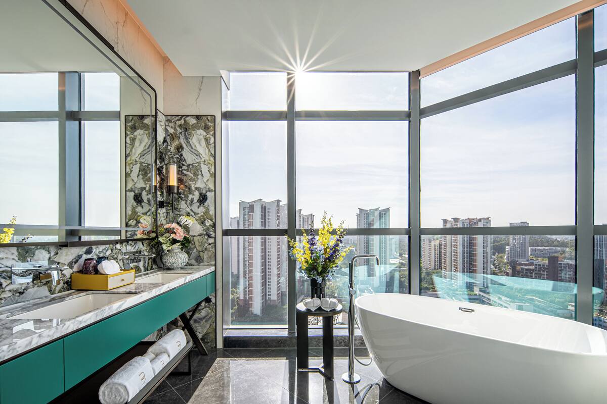 A modern bathroom with a large bathtub, a marble vanity, and a city view through large windows.