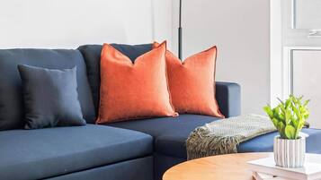 A modern living room with a dark blue sofa, two orange cushions, a small round wooden table, and a potted plant.