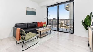 Een moderne woonkamer met een zwarte leren bank, een houten salontafel en een glazen deur naar een buitenbalkon met uitzicht op de stad.