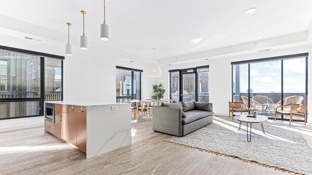 Een ruime woonkamer met een grijze bank, een salontafel en rieten stoelen. Een keuken met een wit aanrecht en koperen kastjes is zichtbaar. Grote ramen bieden veel natuurlijk licht en uitzicht over de stad.