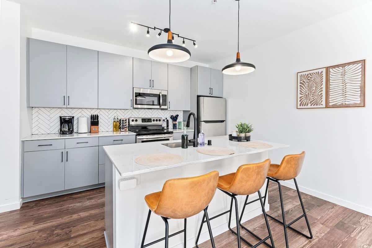 A modern kitchen with grey cabinets, a white island with bar stools, a microwave, a refrigerator, and pendant lighting.