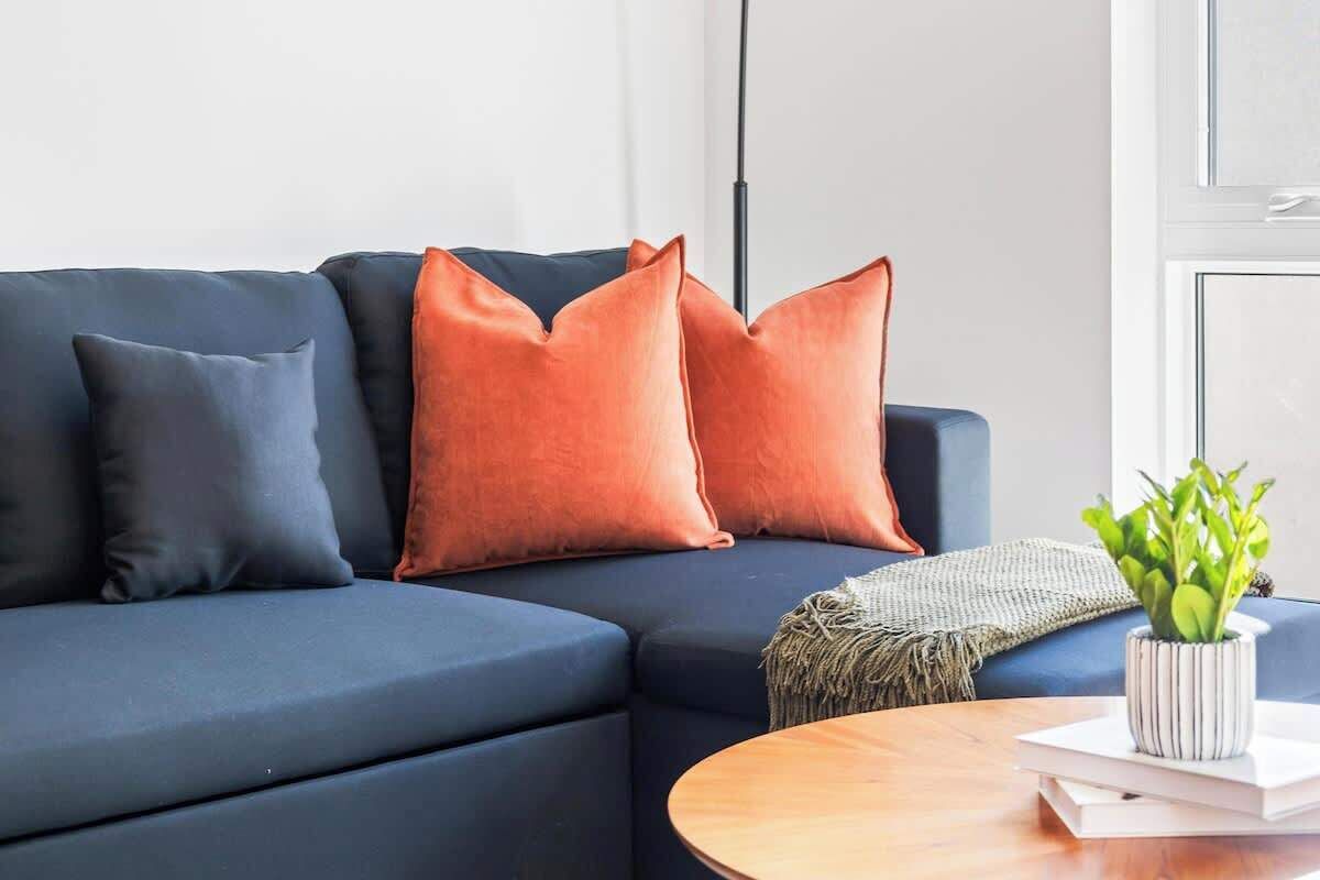 Ein modernes Wohnzimmer mit einem dunkelblauen Sofa, zwei orangefarbenen Kissen, einem kleinen runden Holztisch und einer Topfpflanze.