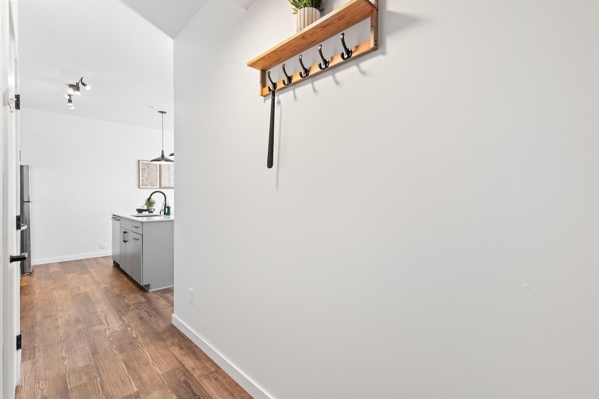 A hallway with a wooden floor, a kitchen area with a sink and cabinets, and a wall-mounted coat rack with hooks.