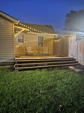 Un patio trasero con terraza de madera, dos sillas, una mesa y luces colgantes.
